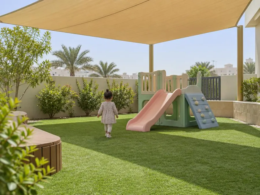 Shaded outdoor play area with artificial grass and a small slide, a toddler seen from behind in the distance